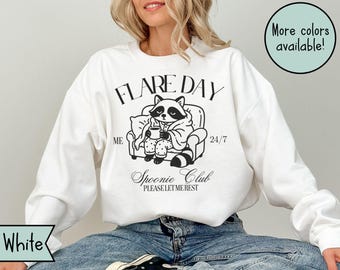 Pode incluir: Camisola branca com um gráfico preto de um guaxinim de pijama, sentado numa cadeira e segurando uma caneca. O texto diz "FLARE DAY" acima do guaxinim e "Spoonie Club" abaixo. O texto adicional diz "ME 24/7" e "PLEASE LET ME REST".