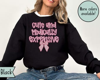 May include: Black sweatshirt with the text "cute and medically expensive" in pink, along with a pink bow graphic. The sweatshirt is worn with blue jeans. A circular graphic in the upper right corner says "More colors available!"