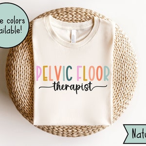 May include: A white t-shirt with the text "PELVIC FLOOR therapist" in rainbow colors. The shirt is on a woven mat with the word "Natural" in the bottom right corner.