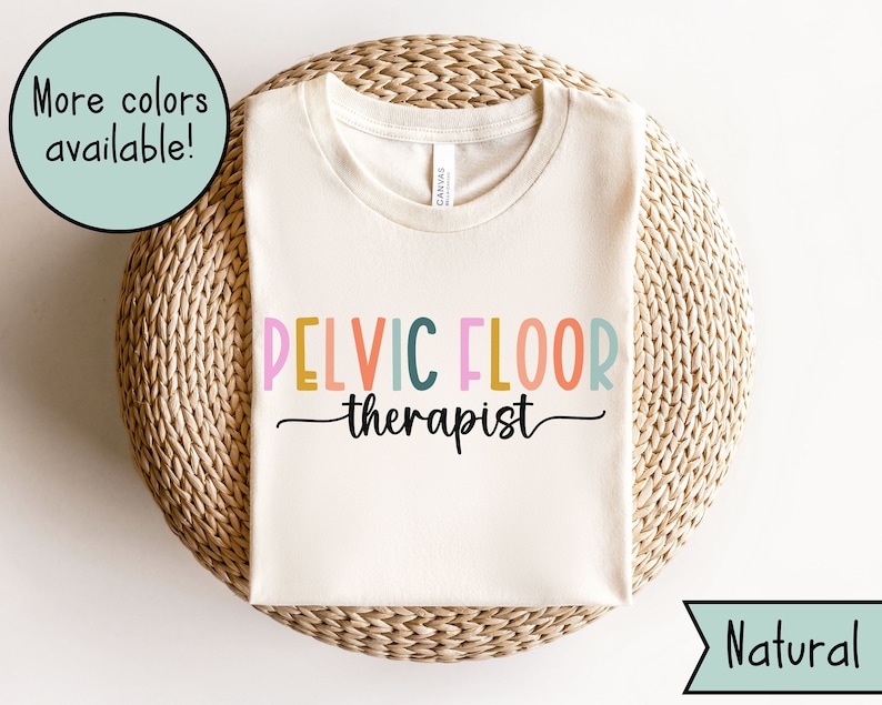 May include: A white t-shirt with the text "PELVIC FLOOR therapist" in rainbow colors. The shirt is on a woven mat with the word "Natural" in the bottom right corner.