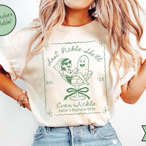 May include: Ivory-colored t-shirt with a green graphic. The graphic features a bride and a pickle in a martini glass, with the text "Last Pickle She'll Ever Fickle Katie's Bachelorette" and the year '25.