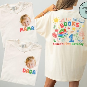 May include: Off-white t-shirts featuring a child's photo and colorful text. The back design includes the text 'ONE FOR THE BOOKS' and 'Emma's First Birthday' with illustrations of books, a caterpillar, a balloon, a gift, and a cake.