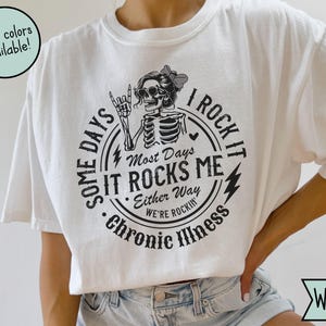 Könnte beinhalten: Weißes T-Shirt mit schwarzer Grafik, die ein Skelett mit Sonnenbrille und Schleife zeigt, umgeben von dem Text "Some days it rocks me, I rock it, chronic illness." Weitere Farben sind verfügbar.