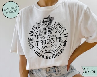 Może przedstawiać: Biała koszulka z czarną grafiką przedstawiającą szkielet w okularach przeciwsłonecznych i kokardą, otoczony tekstem "Some days it rocks me, I rock it, chronic illness." Dostępne są inne kolory.