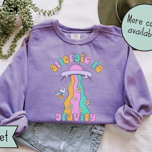 May include: A purple sweatshirt with a graphic of a cartoon spaceship with a rainbow trail and the text "allergic to gravity".