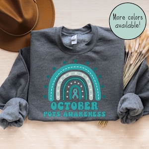 May include: A dark grey sweatshirt featuring a teal blue and white rainbow design. The rainbow has a teal blue ribbon in the centre and the text "OCTOBER POTS AWARENESS" below it. The text "More colours available!" is in a teal blue circle in the top right corner of the image.