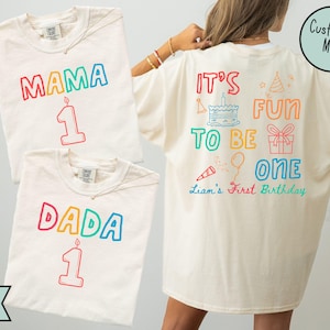 May include: Ivory t-shirts with colourful text and graphics. One shirt says "MAMA 1" and another says "DADA 1". The back of a third shirt reads "IT'S FUN TO BE ONE" with birthday-themed illustrations and "Liam's First Birthday".