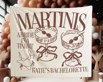 Könnte beinhalten: Ein beigefarbener Stoffbanner mit braunem Text, der "Martini's Abride Ina Tini Bit Katie's Bachelorette" lautet. Der Banner zeigt zwei Illustrationen von Martini-Gläsern mit Oliven.