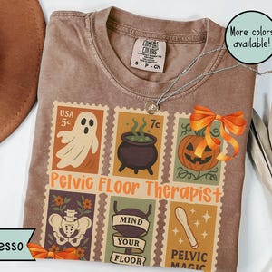 May include: Espresso-colored t-shirt with a stamp-themed design featuring a ghost, cauldron, jack-o'-lantern, and other graphics. The text "Pelvic Floor Therapist" is displayed. A silver necklace and an orange bow are also visible.