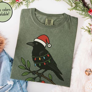 May include: Olive green t-shirt featuring a black crow wearing a Santa hat and string lights. The crow is perched on a branch with green leaves and red berries. The text "More colors available!" is in a circle.