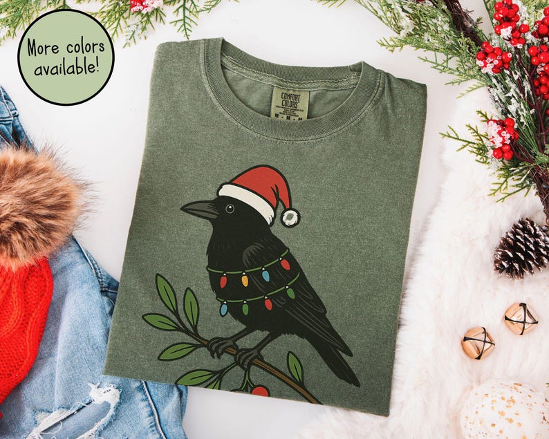 May include: Olive green t-shirt featuring a black crow wearing a Santa hat and string lights. The crow is perched on a branch with green leaves and red berries. The text "More colors available!" is in a circle.