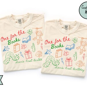 May include: Two ivory-colored t-shirts with the text "One for the Books" in red and green, along with colorful illustrations of books, a monkey, and a caterpillar. The text "Liam's First Birthday" is also present.