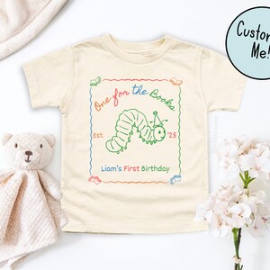 May include: Cream-colored toddler t-shirt with a green caterpillar graphic and the text "One for the Books." The design includes a red, green, and blue border, with the text "Liam's First Birthday." A teddy bear and flowers are in the background.