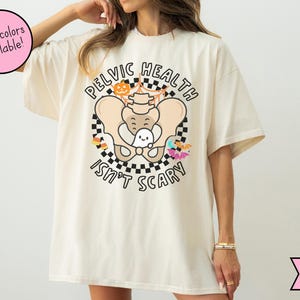May include: Ivory t-shirt featuring the text "PELVIC HEALTH ISN'T SCARY" with a cartoon pelvis, ghost, and Halloween decorations. The shirt has a loose fit. More colours are available.