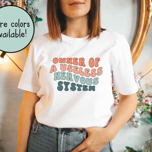 May include: White t-shirt with a colorful graphic that reads "OWNER OF A USELESS NERVOUS SYSTEM".