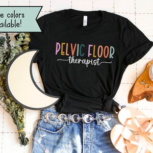 May include: Black t-shirt with the text "PELVIC FLOOR therapist" in colorful letters. The shirt is laid out on a white surface with a pair of sandals, a belt, and a cup of coffee.