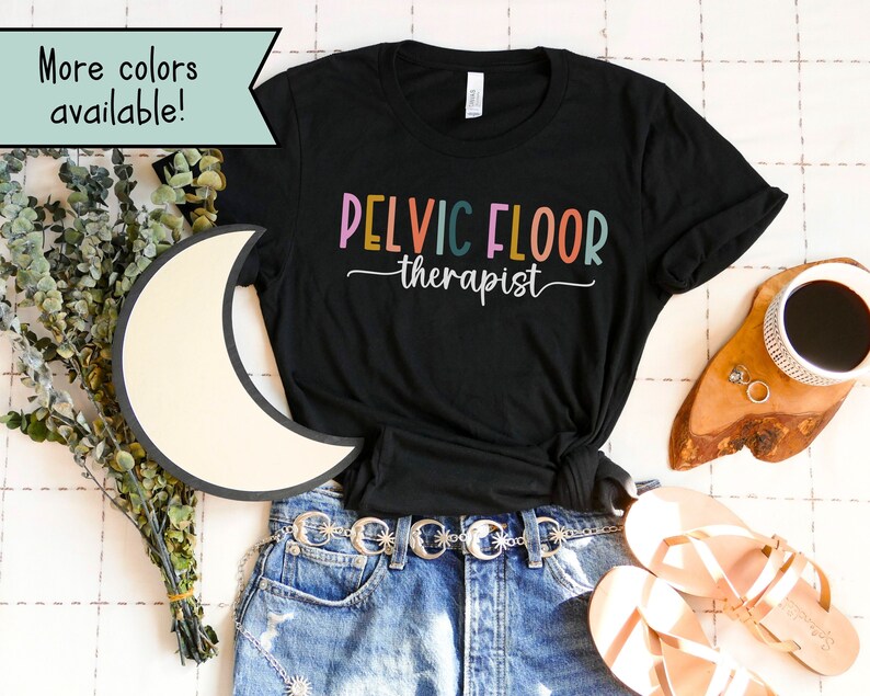 May include: Black t-shirt with the text "PELVIC FLOOR therapist" in colorful letters. The shirt is laid out on a white surface with a pair of sandals, a belt, and a cup of coffee.