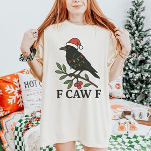 May include: Cream-colored t-shirt featuring a black crow wearing a Santa hat and Christmas lights, perched on a branch with red berries. The text "F CAW F" is printed below the graphic. The shirt is part of a holiday-themed apparel collection.