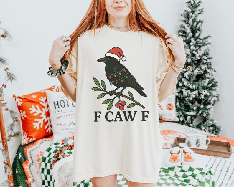May include: Cream-colored t-shirt featuring a black crow wearing a Santa hat and Christmas lights, perched on a branch with red berries. The text "F CAW F" is printed below the graphic. The shirt is part of a holiday-themed apparel collection.