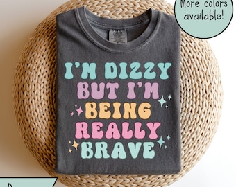 Op de afbeelding: Een grijs T-shirt met de tekst "I'm dizzy but I'm being really brave" in kleurrijke retro-lettertypen. De tekst is omringd door kleine gele sterren.