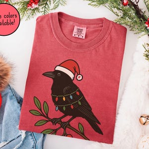 May include: Red t-shirt featuring a black crow wearing a Santa hat and string lights. The crow is perched on a branch with green leaves. Text in a pink circle says "More colors available!"