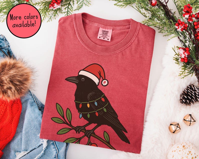 May include: Red t-shirt featuring a black crow wearing a Santa hat and string lights. The crow is perched on a branch with green leaves. Text in a pink circle says "More colors available!"