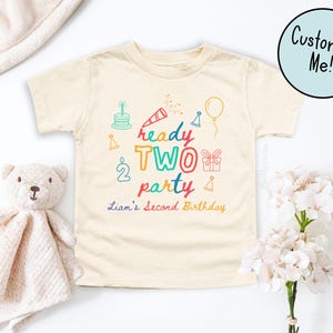 Può includere: Una maglietta color crema per bambini piccoli con una grafica colorata che recita "ready TWO party" con illustrazioni a tema compleanno. Il testo "Liam's Second Birthday" è in basso. Un orsacchiotto e dei fiori sono nell'immagine.