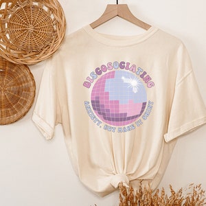 May include: A cream-colored t-shirt with a graphic of a disco ball and the text "Discosociating Anxiety, But Make It Shiny".