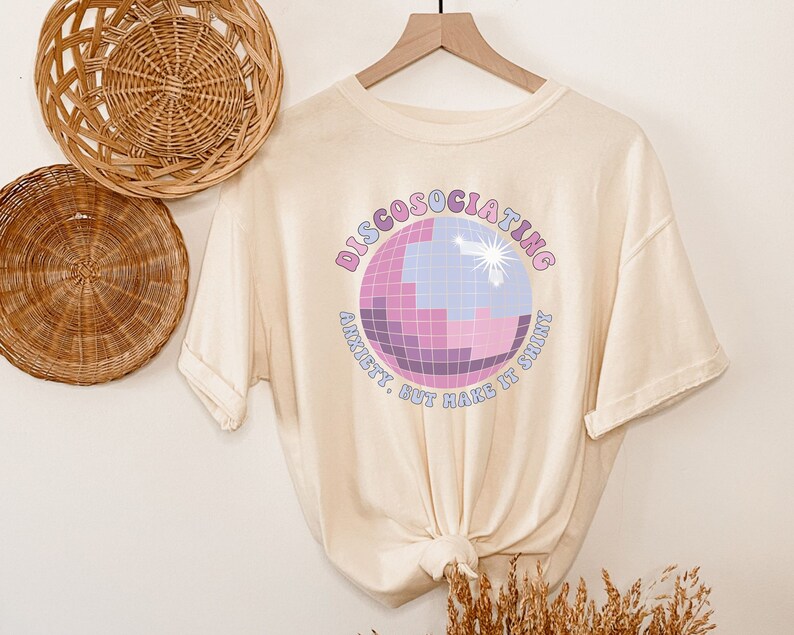 May include: A cream-colored t-shirt with a graphic of a disco ball and the text "Discosociating Anxiety, But Make It Shiny".