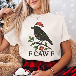 May include: Cream-colored t-shirt featuring a black crow wearing a Santa hat and string lights, perched on a branch with berries. The text "F CAW F" is printed below the illustration. The shirt is part of a holiday collection.