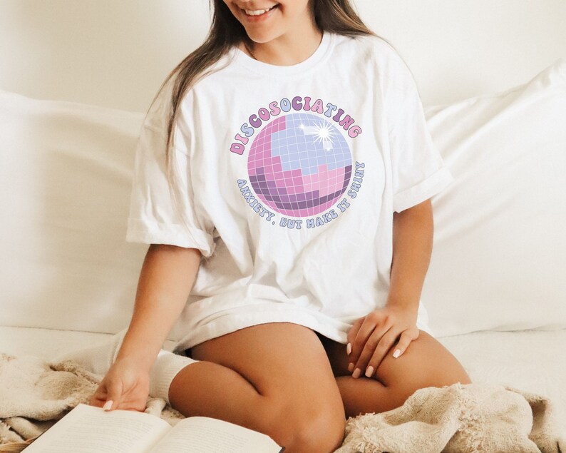 May include: White t-shirt with a pink and purple disco ball graphic and the text "Discosociating. Anxiety. But make it shiny."