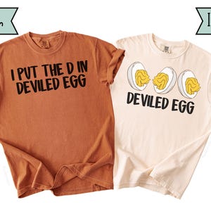 May include: Two t-shirts are shown, one in a yam colour and the other in ivory. The yam-coloured shirt has the text "I PUT THE D IN DEVILED EGG" in black. The ivory shirt features a graphic of three devilled eggs and the text "DEVILED EGG".
