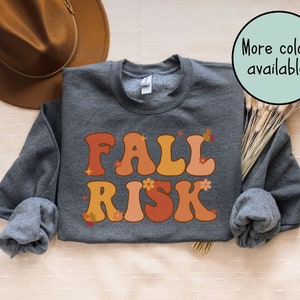 May include: A dark grey sweatshirt featuring the text "FALL RISK" in orange and peach bubble letters, decorated with small flower and leaf motifs. A green circle displays the text "More colours available!"