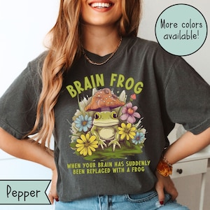 May include: A dark grey t-shirt with a green frog wearing a mushroom hat. The shirt has the text "BRAIN FROG" in green and yellow letters and the text "WHEN YOUR BRAIN HAS SUDDENLY BEEN REPLACED WITH A FROG" in black letters.