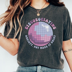 May include: A dark gray t-shirt with a disco ball graphic and the text "DISCOSOCIATING ANXIETY BUT MAKE IT SHINY".