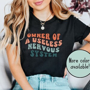 May include: A black t-shirt with the text "Owner of a useless nervous system" in a retro font with a gradient of red, orange, and blue. The text is centered on the shirt. The shirt is being worn by a person with long blonde hair. The text "More colors available!" is in a circle on the bottom right of the image.