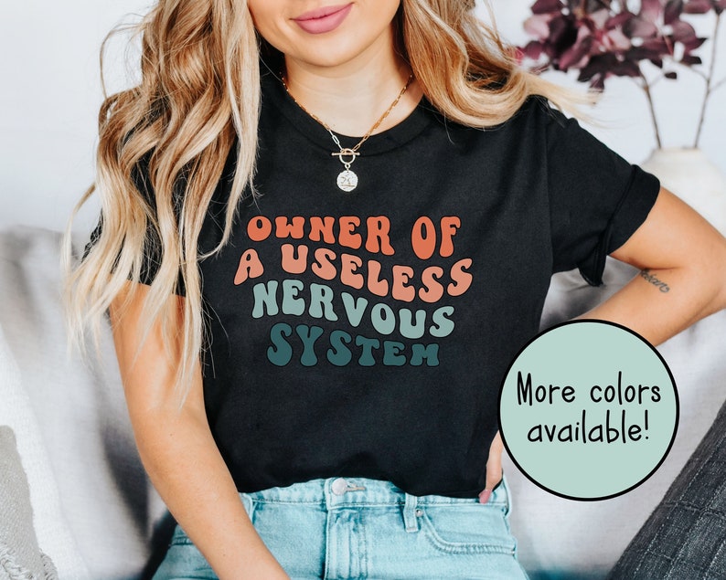 May include: A black t-shirt with the text "Owner of a useless nervous system" in a retro font with a gradient of red, orange, and blue. The text is centered on the shirt. The shirt is being worn by a person with long blonde hair. The text "More colors available!" is in a circle on the bottom right of the image.
