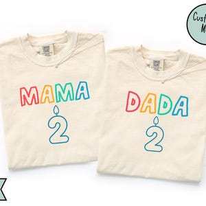 May include: Ivory-colored t-shirts with the words "MAMA" and "DADA" in rainbow-colored bubble letters, each with a number "2" and a candle graphic. The shirts have a round neckline and short sleeves. The text "Customize Me!" is in a circle.