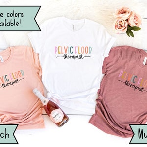 May include: Three t-shirts in peach, white, and mauve colors with the text "PELVIC FLOOR therapist" printed on them. The shirts are displayed on a white wooden surface.