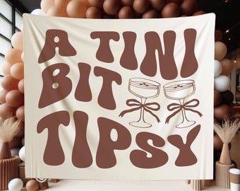 Könnte beinhalten: Ein weißer Wandteppich mit braunem Text, der "A TINI BIT TIPSY" lautet. Zwei braune Cocktailgläser mit braunen Stielen befinden sich auf beiden Seiten des Textes.