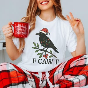 May include: White t-shirt featuring a cartoon crow wearing a Santa hat and Christmas lights, perched on a branch with berries. The text "F CAW F" is below the crow. A red mug with "Have yourself a Merry Little Christmas" is held in the hand.