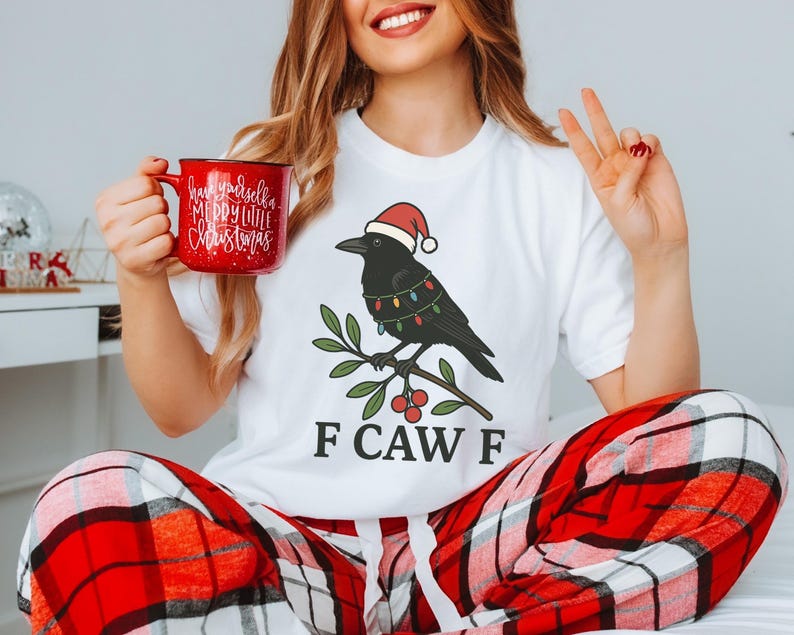 May include: White t-shirt featuring a cartoon crow wearing a Santa hat and Christmas lights, perched on a branch with berries. The text "F CAW F" is below the crow. A red mug with "Have yourself a Merry Little Christmas" is held in the hand.