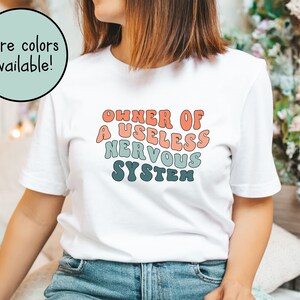 May include: White t-shirt with a retro-style graphic that says "Owner of a Useless Nervous System" in a rainbow color scheme.