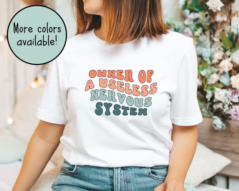 May include: White t-shirt with a retro-style graphic that says "Owner of a Useless Nervous System" in a rainbow color scheme.