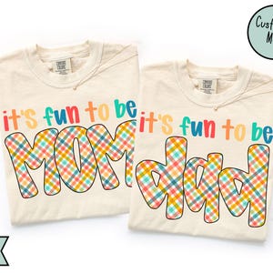 May include: Two ivory t-shirts with the text "it's fun to be" in colourful letters. One shirt says "MOM" and the other says "DAD" in a plaid pattern. A circular graphic says "Customize Me!"