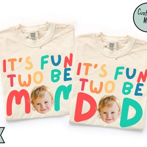 May include: Two ivory-colored t-shirts with the text "IT'S FUN TWO BE MOM/DAD" in colorful letters. Each shirt features a photo of a child's face. The shirts have a round neckline and short sleeves. A gold necklace is visible on each shirt.