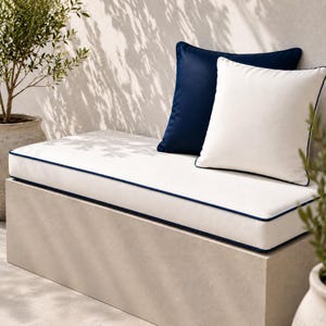 May include: A rectangular white bench cushion with dark blue piping along the edges, accompanied by one solid dark blue square throw pillow and one solid white square throw pillow with dark blue piping, arranged on a light beige stone base.
