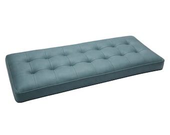 Puede incluir: Un cojín rectangular para banco en color azul cerceta apagado. El cojín presenta una superficie capitoné con múltiples botones empotrados dispuestos en cuadrícula, creando una serie de hendiduras suaves y redondeadas.
