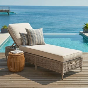 May include: A beige cushioned chaise lounge with a woven gray frame and two striped pillows. A small round wicker side table holds a tray with a glass. The scene is set by a pool and the ocean.