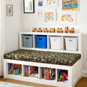 May include: A white storage bench with a leopard print cushion, cubby shelves, and colorful books. The top shelf holds gray, blue, and white storage bins, and wooden toy trains. Artwork hangs on the white wall.